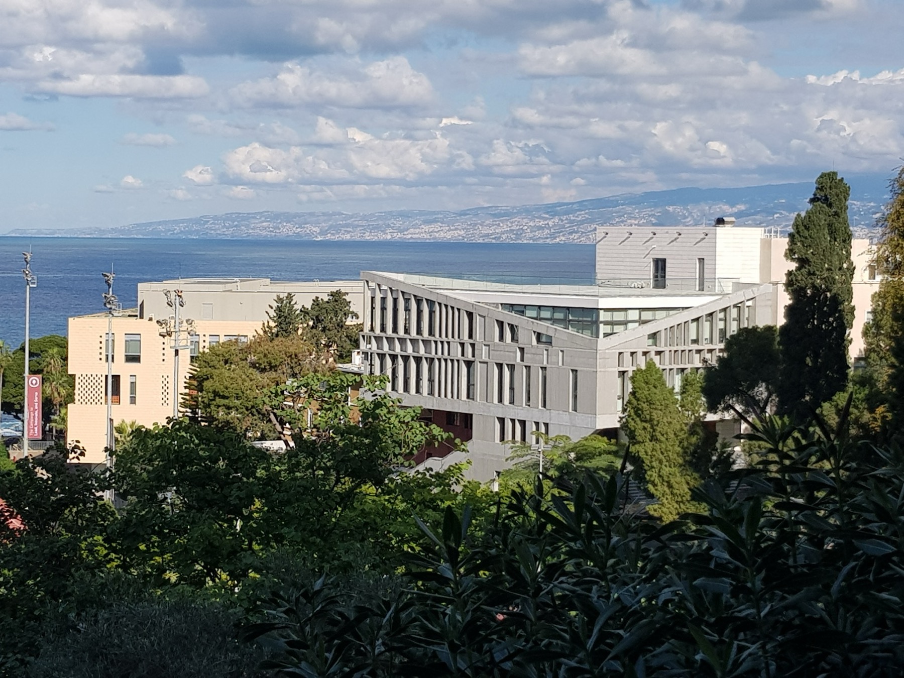 A modern grey building with an angled roof among trees on the AUB campus, with the Mediterranean Sea and the coast behind it.