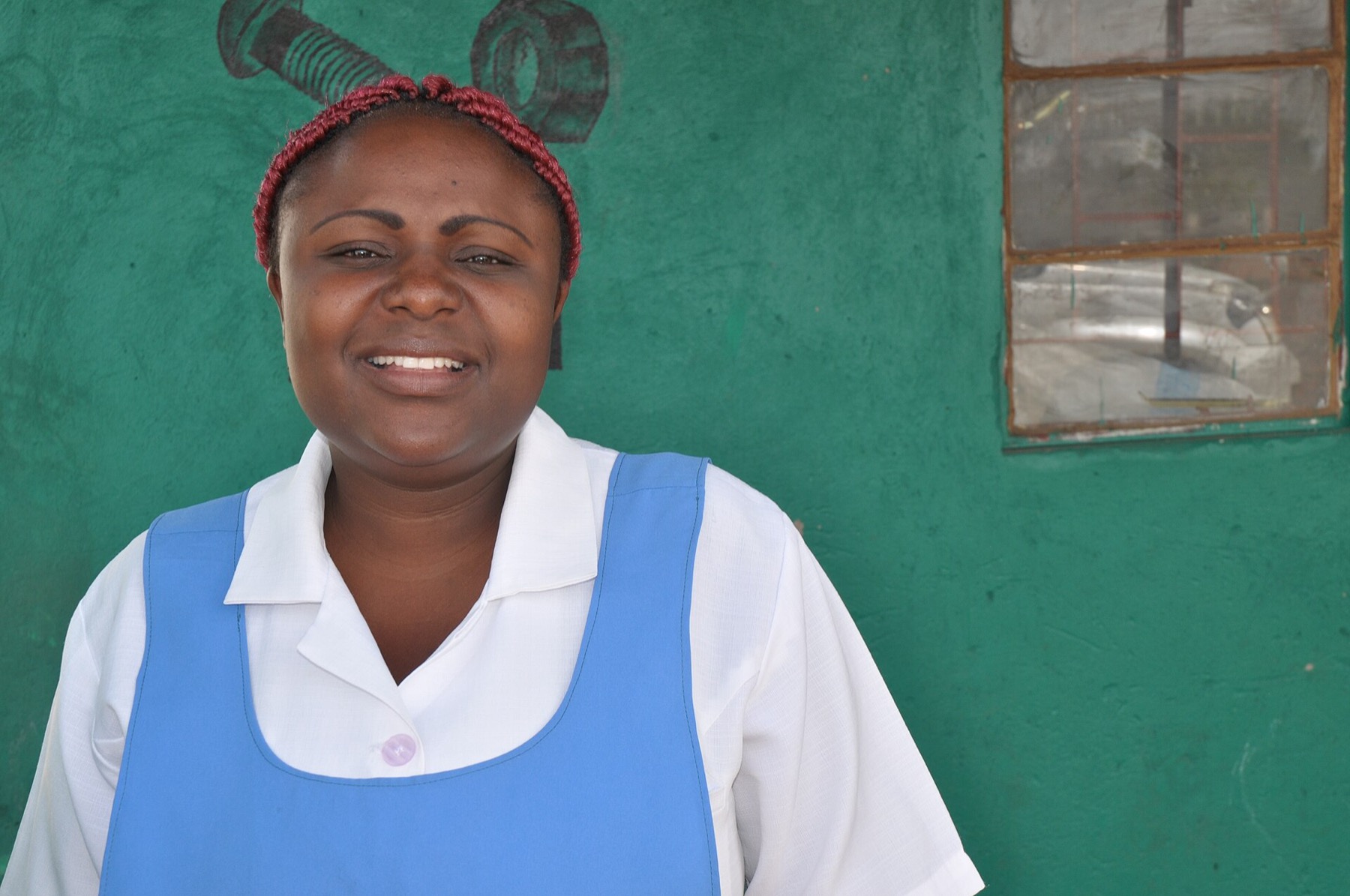 A smiling nurse midwife in a white uniform and blue pinafore stands in front of a green wall at a rural health center in Zambia