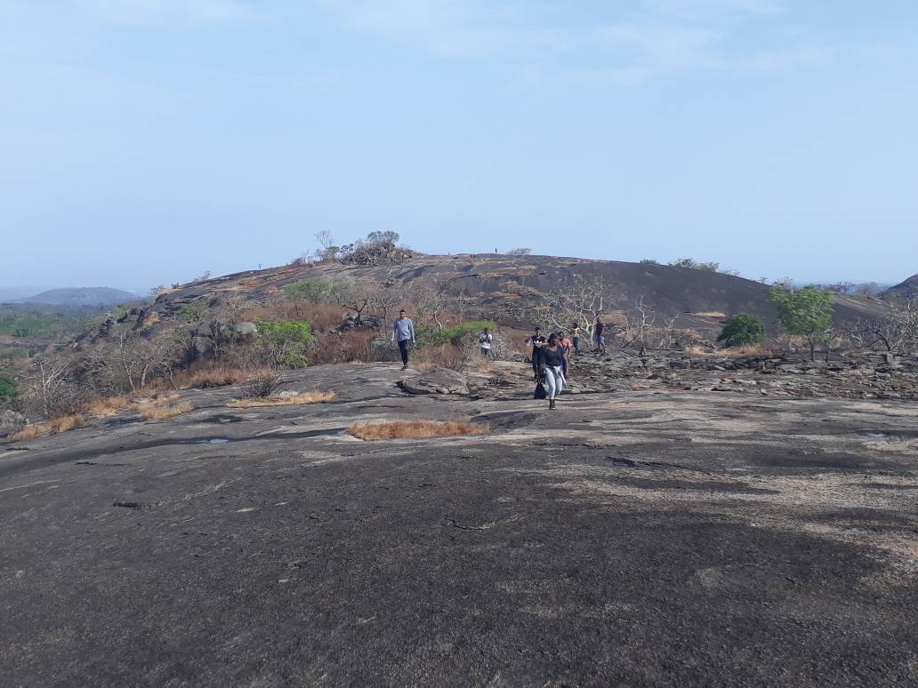 A wide dark granite rock with dry grass and bare shrubs under a hazy blue sky, with a few small hikers in the distance