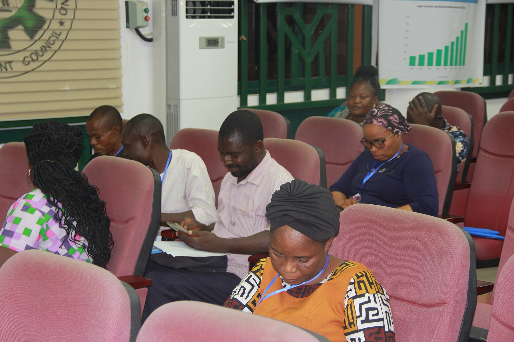 Adult teachers sit in rows of pink auditorium chairs, most looking at phones or papers, during a training session in Abuja.