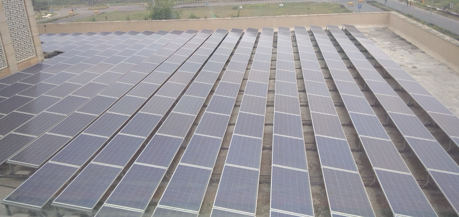 Rows of tilted blue solar panels covering a flat concrete roof, with a road and trees beyond.